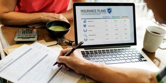 A person reviewing insurance plans on a laptop while filling out paperwork at a table with a bowl and calculator.