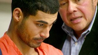 A young man in an orange jumpsuit appears attentive as another individual speaks to him, likely in a courtroom setting.
