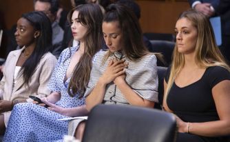 Four gymnasts sitting in a Senate hearing audience, conveying emotions of concern and contemplation.