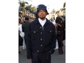 Person wearing a navy jacket, white shirt, and blue bucket hat at an outdoor event with a crowd and camera crews in the background.