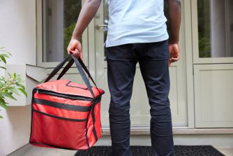 A delivery worker holding a red insulated food delivery bag standing at a residential doorway.