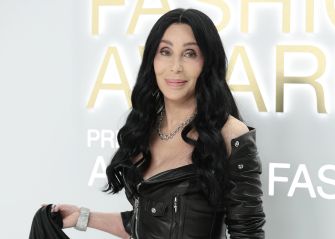 Close-up portrait of a woman with long black hair wearing a black leather jacket and silver jewelry, posing in front of a backdrop with large gold letters.