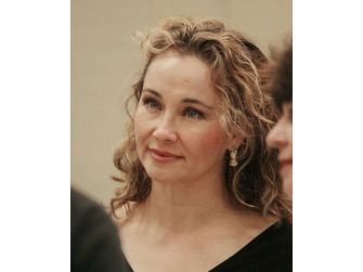 Close-up portrait of a woman with light blonde, wavy hair and blue eyes, wearing dangling earrings.