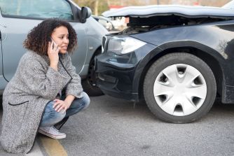 Woman sitting on the ground beside a damaged car, talking on her cell phone after a traffic accident.