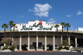 Front view of the Rose Bowl stadium entrance with a large "Rose Bowl" sign, tall columns, palm trees, and a blue sky.