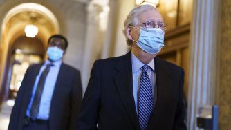 Two men in suits walking through a corridor, one wearing a mask.