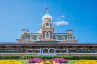 Front facade of Magic Kingdom entrance with clock tower, American flag, and colorful flower beds under a blue sky.