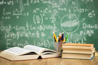 A classroom setting featuring an open book, stacked books, and a cup of colorful pencils in front of a green chalkboard with mathematical equations.