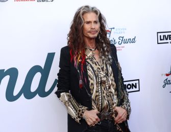 Person with long, wavy hair wearing a patterned shirt and layered jewelry, posing in front of a white backdrop with logos.