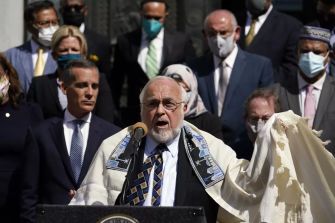 A group of individuals, including a speaker in traditional Jewish attire, participate in a rally addressing rising antisemitism and hate crimes against the Jewish community.