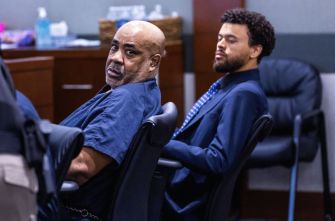Courtroom scene with two men in blue jail jumpsuits seated; the bald man in the foreground looks toward the camera, while the younger man with curly hair sits behind him.