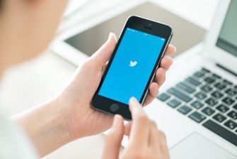 A person holding a smartphone displaying the Twitter logo on the screen, with a laptop visible in the background.