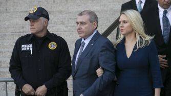 Two individuals, flanked by a police officer, leaving a courtroom following a hearing related to fraud charges.