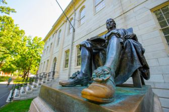 Statue of a seated figure in front of a university building, surrounded by greenery.