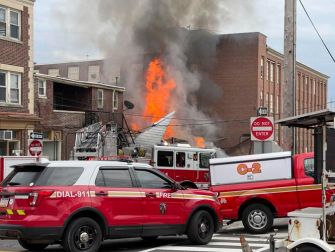 Fire and smoke erupting from a building after an explosion, with emergency vehicles and firefighters responding on the scene.