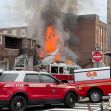 Fire and smoke erupting from a building after an explosion, with emergency vehicles and firefighters responding on the scene.