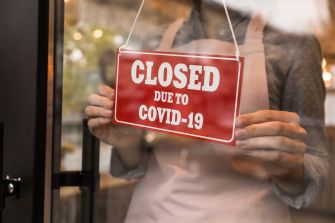 A person holding a red "CLOSED DUE TO COVID-19" sign in front of a shop window.
