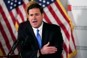 Governor Doug Ducey of Arizona speaking at a press conference, with American flags in the background.
