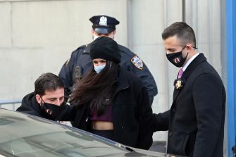 People wearing masks outside a building near a parked car, with two uniformed officers in the background.