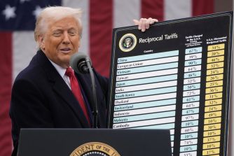 A man in a dark suit and red tie speaks at a podium, holding a large chart labeled "Reciprocal Tariffs" with country names and percentage columns, the American flag in the background and the presidential seal on the podium.