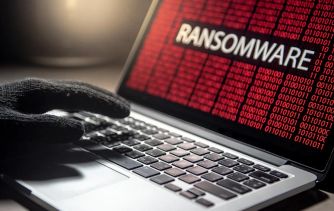 A hand wearing a black glove on a laptop keyboard, with a screen displaying the word "RANSOMWARE" in bold letters against a red background.