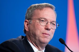 A man with glasses speaks into a microphone against a blue background.