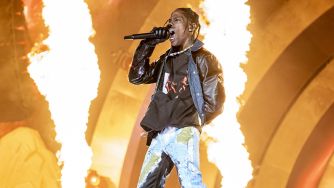 Travis Scott performing on stage with flames in the background during a concert.