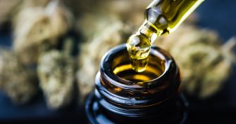 A drop of cannabis oil being dispensed from a glass dropper into a small amber bottle, with cannabis buds visible in the background.