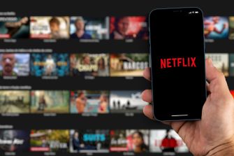 A hand holding a smartphone displaying the Netflix logo, with a blurred Netflix streaming library in the background.