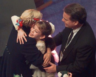 A figure skater in a sparkly dress is embraced by a coach while a man in a suit watches.
