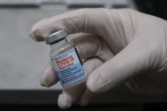 A gloved hand holding a vial of Moderna COVID-19 vaccine.