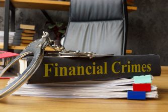 A close-up of a book titled "Financial Crimes" with handcuffs placed on top, surrounded by papers in an office setting.