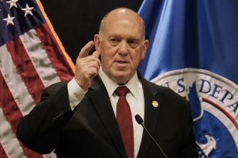 A man in a dark suit and red tie speaks at a podium, gesturing with his finger near his temple, with the American flag and a blue banner in the background.