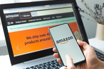 A person holding a smartphone displaying the Amazon logo, with an Amazon shopping website open on a laptop in the background.