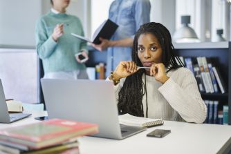 A woman with dreadlocks sitting at a desk with a laptop, looking directly at the camera while two other individuals converse in the background.
