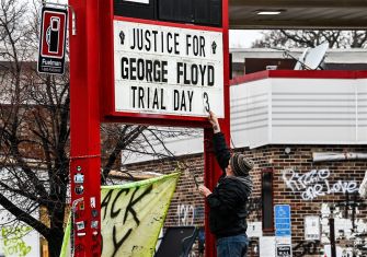 A person hangs a sign reading JUSTICE FOR GEORGE FLOYD TRIAL DAY on a red street pole, with graffiti and a storefront in the background.