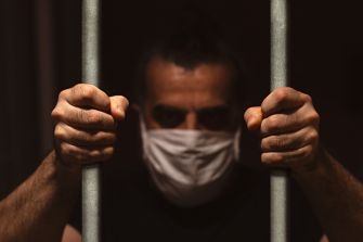 A man wearing a mask grips prison bars, highlighting the struggles of inmates during the COVID-19 pandemic.