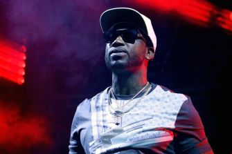 Close-up of a male rapper wearing a white cap, dark sunglasses, and chains, performing on a stage with red lighting.
