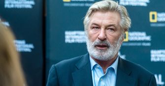 Alec Baldwin speaking at an event, with a focused expression amid a backdrop featuring a logo.