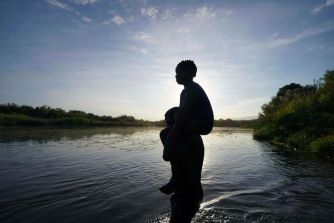 A person carries a child on their shoulders while crossing a river, with a picturesque landscape in the background.