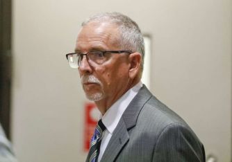 A man in a suit and glasses stands in a hallway, appearing serious and focused.