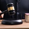 Close-up of a judge's gavel about to strike a sound block on a wooden table, with a law book nearby.