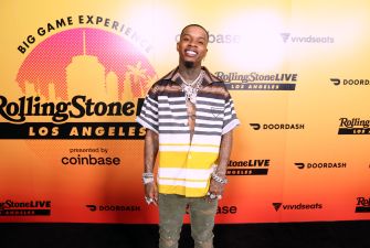 Man with tattoos wearing a striped yellow and white shirt and heavy jewelry, posing in front of a Rolling Stone Live Los Angeles backdrop with sponsor logos.