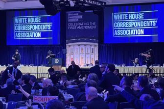 Blue-lit stage for the White House Correspondents’ Association dinner with a podium and large screens displaying the WHCA logo and “Celebrating the First Amendment,” surrounded by photographers and seated guests.