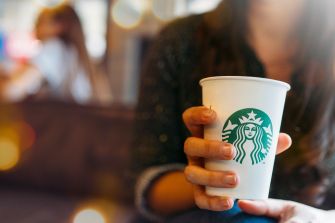 Close-up of a hand holding a white Starbucks cup with the green siren logo, blurred cafe background.