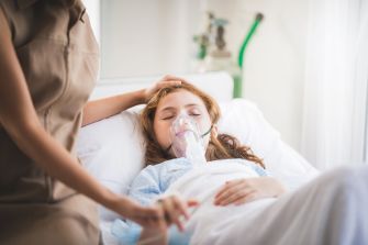 A young girl lies in a hospital bed with an oxygen mask, cared for by an adult.