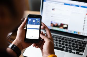 A person using a smartphone to access Facebook, with a laptop open in the background displaying the social media platform.