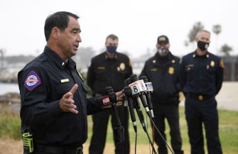 Uniformed law enforcement officers hold a press briefing on a beach, with microphones set up in front of them.