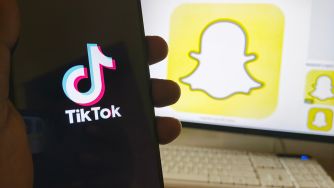 Close-up of a smartphone displaying the TikTok logo, held by a hand, with a large Snapchat ghost logo on a computer screen in the background.