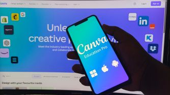 Close-up of a hand holding a smartphone displaying the Canvas Education Pro app, with a computer monitor in the background showing the Canvas branding.